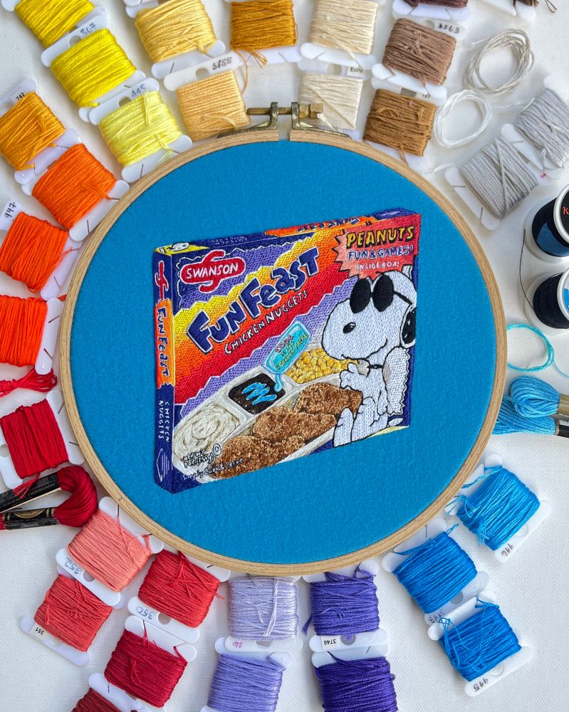 Embroidery of a 90s Cool Snoopy Fun Feast TV dinner with mashed potatoes, corn, chicken nuggets and a brownie being coated in disturbingly blue icing from a floating “squeeze packet.”