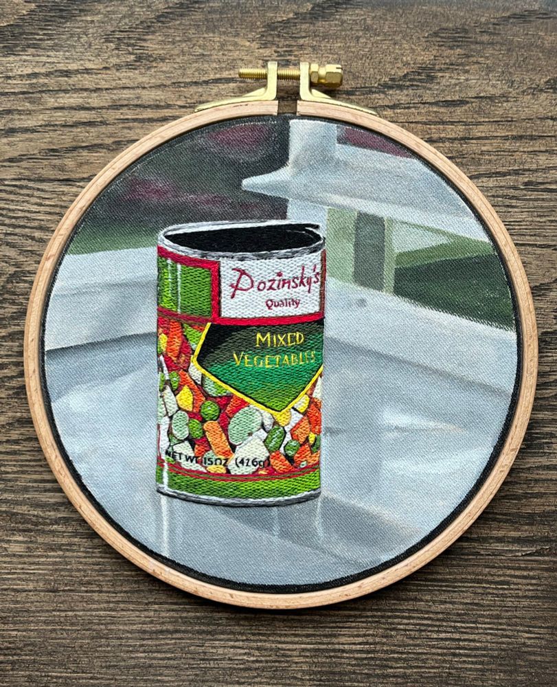 An embroidered can of vegetables on a painted background of a metal tray in a wooden embroidery hoop.