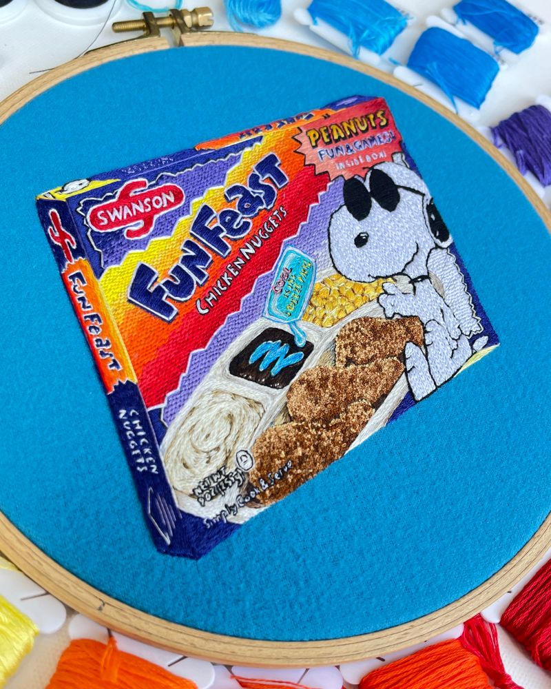 Slight angle of my embroidery of a 90s Cool Snoopy Fun Feast TV dinner with mashed potatoes, corn, chicken nuggets and a brownie being coated in disturbingly blue icing from a floating “squeeze packet.”
