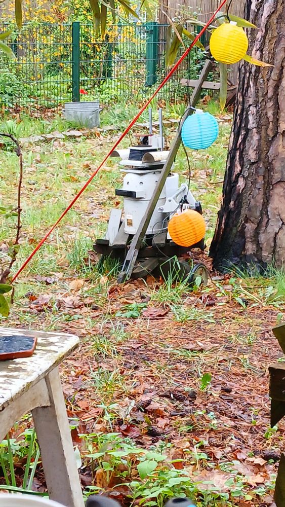 In einem Garten steht ein verrottender Bollerwagen, darauf ein alter Belichtungsapperat. Das Gerät sieht aus, wie ein Vogel mit einem metallhelm oder ein postapokalypsemaulwurf im metallanzug mit komischen Hut. Im Vordergrund bunter Lampions einer Lichterkette.