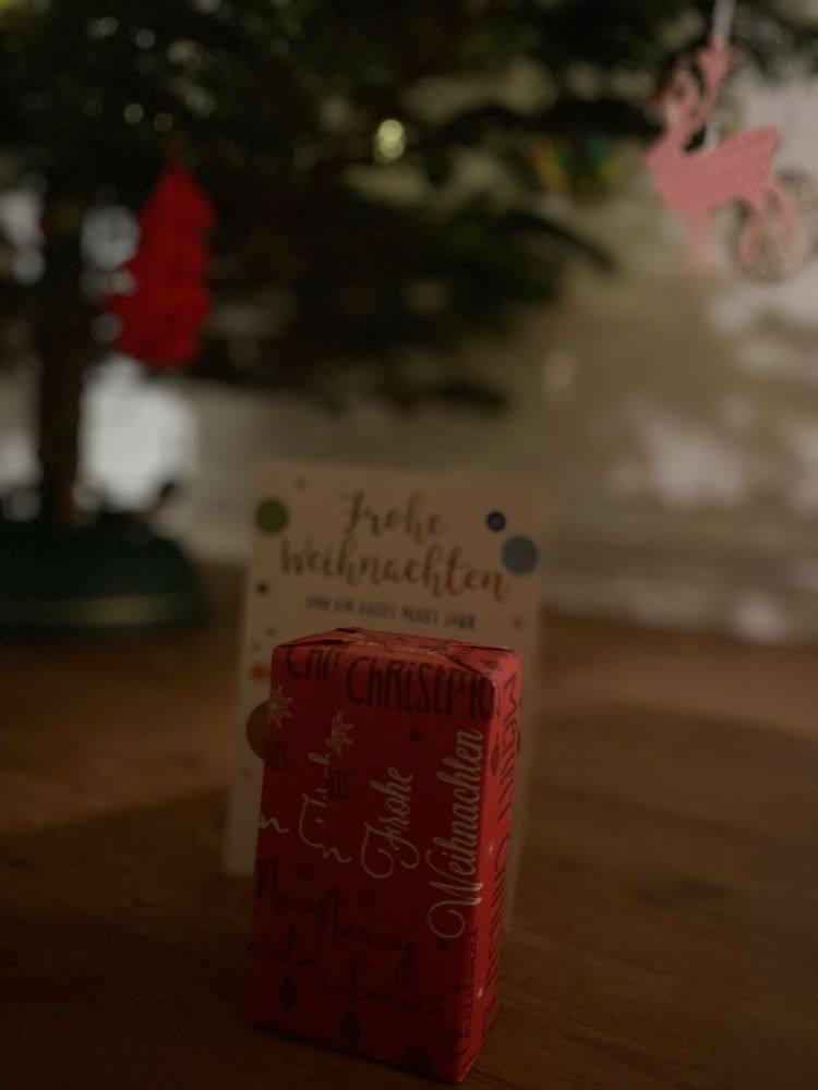Rotes kleines Geschenk mit Weihnachtskarte dahinter vor einem Weihnachtsbaum stehend