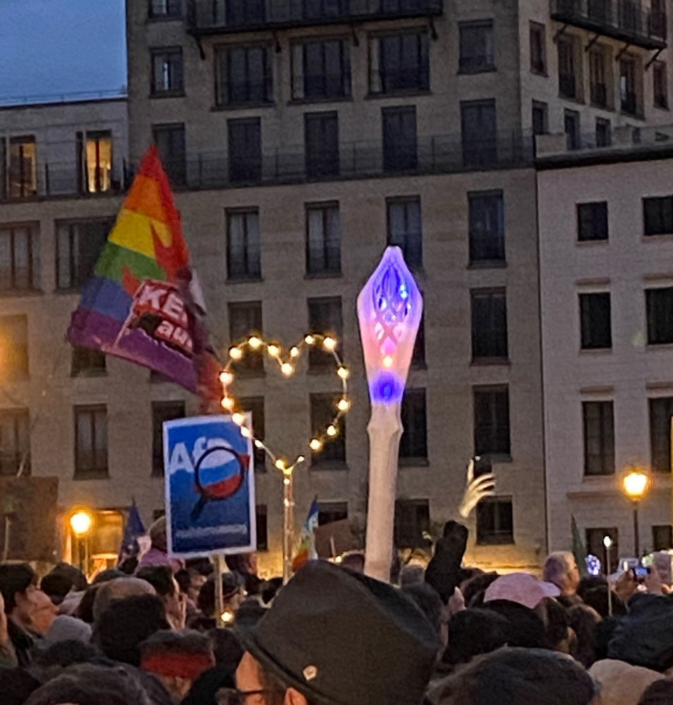 Ein Herz aus einer Lichterkette an einer Stange, ein Leuchtstab und Fahnen und ein Plakat getragen von Teilnehmern und Teilnehmerinnen der Demo