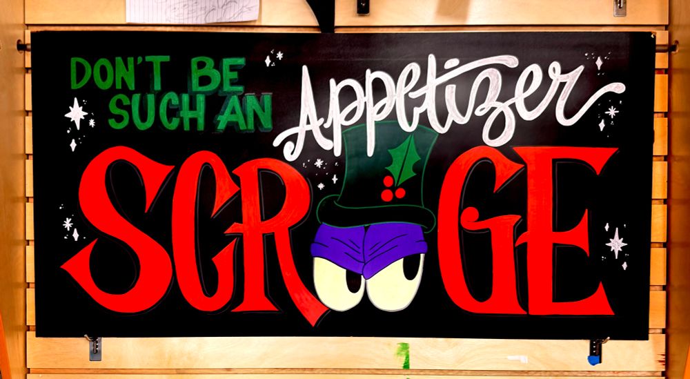 Chalkboard display sign with decorative font saying “Don’t Be Such an Appetizer Scrooge” with two mean looking eyes with a hat on top of them to replace the two “oo” in Scrooge.