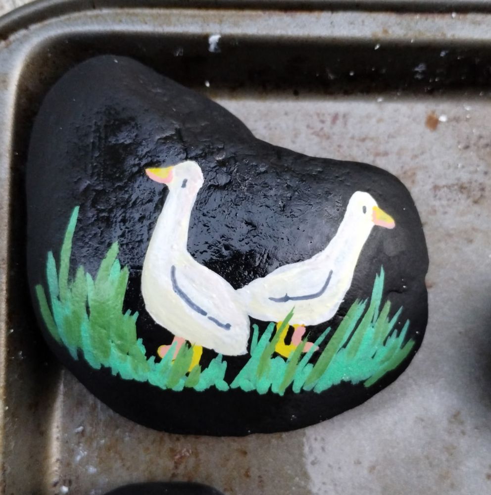 A photo of a rock painted in black that has a scene of two ducks standing in tall grass on it. The paint is acrylic and details are done in paint markers. The ducks are both white with yellow beaks and feet!
