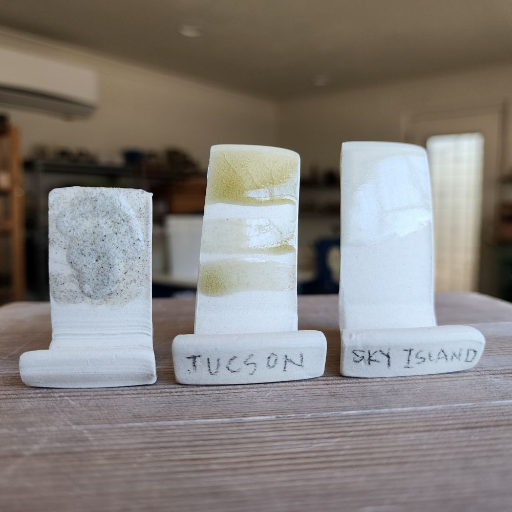 Three porcelain test tiles in the foreground. Left to right: The left tile is a gritty primitive attempt at making a rock glaze using local materials. The middle tile is another attempt using local materials that created a tan, cracking glaze. The right tile is finally looking like a clear glaze! Yay! 