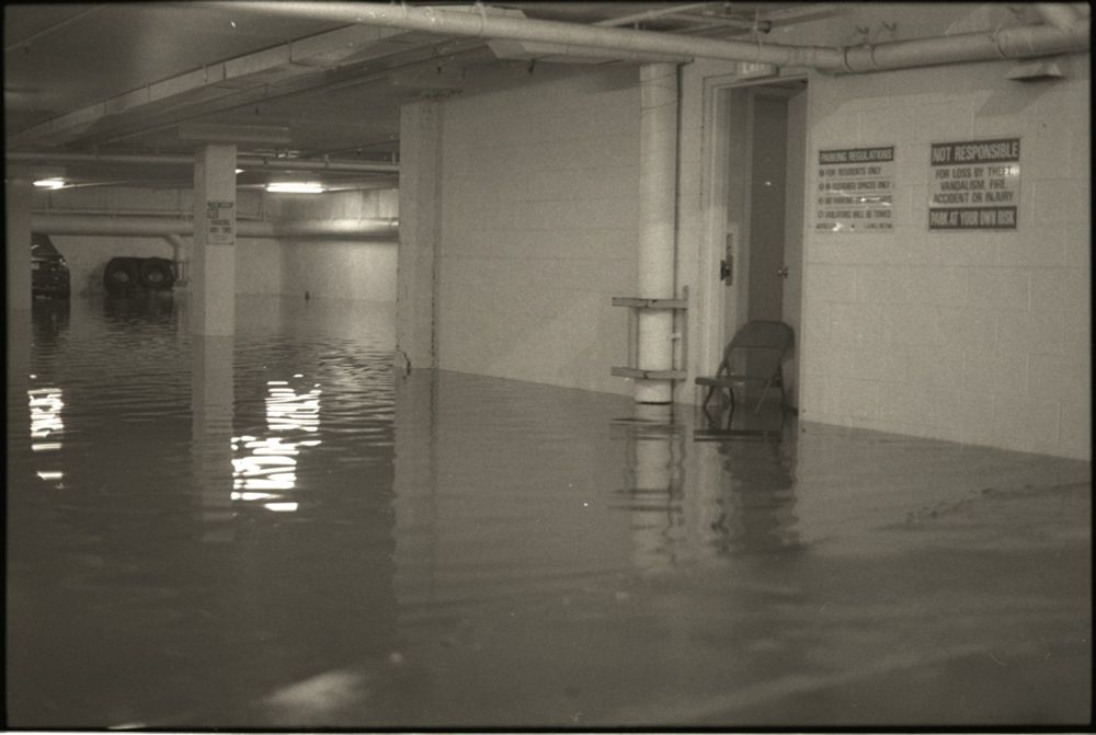 black and white photograph of a flooded garage.  a single metal folding chair props open a door.  
