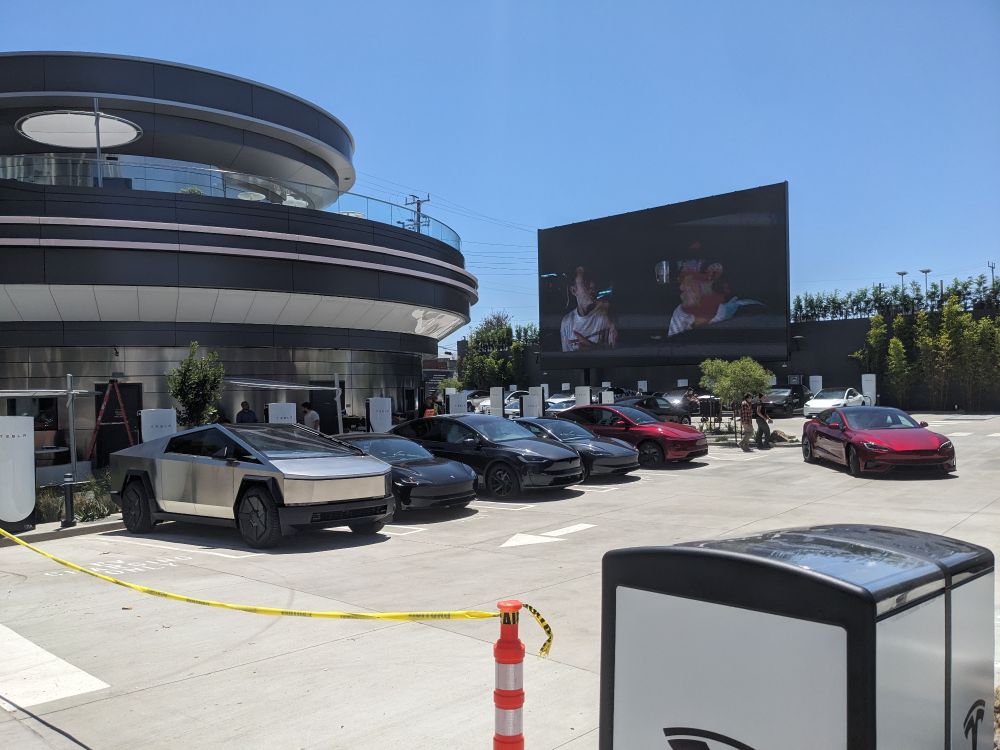 Tesla Diner parking lot with various models of Tesla vehicles including a cybertruck.  A Tesla branded recycle and trash receptacle in the foreground and a jumbotron playing "American Graffiti" in the background. Cautoon tape blocks the driveway entrance.