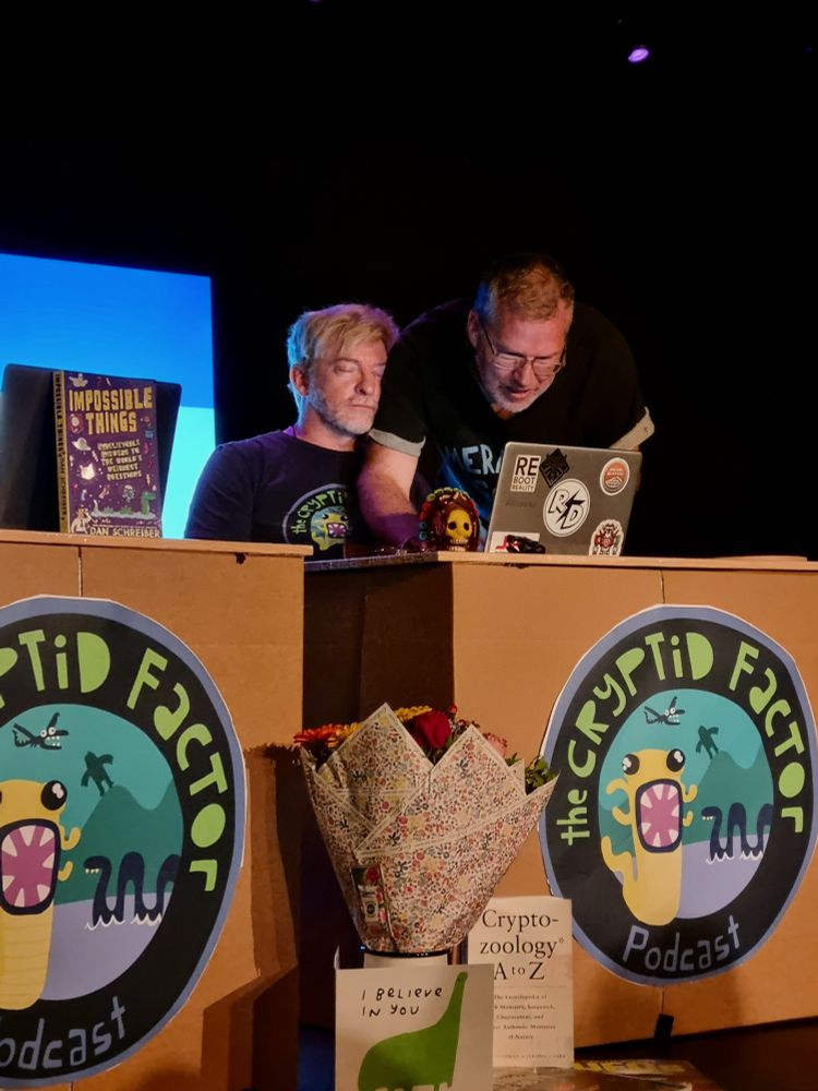 rhys resting his head on buttons's shoulder at the cryptid factor edinburgh fringe show last year. his eyes are closed and he looks content, like he's taking a little snooze while buttons looks for something on rhys's laptop. their desks are cardboard boxes with the cryptid factor logo stuck onto them