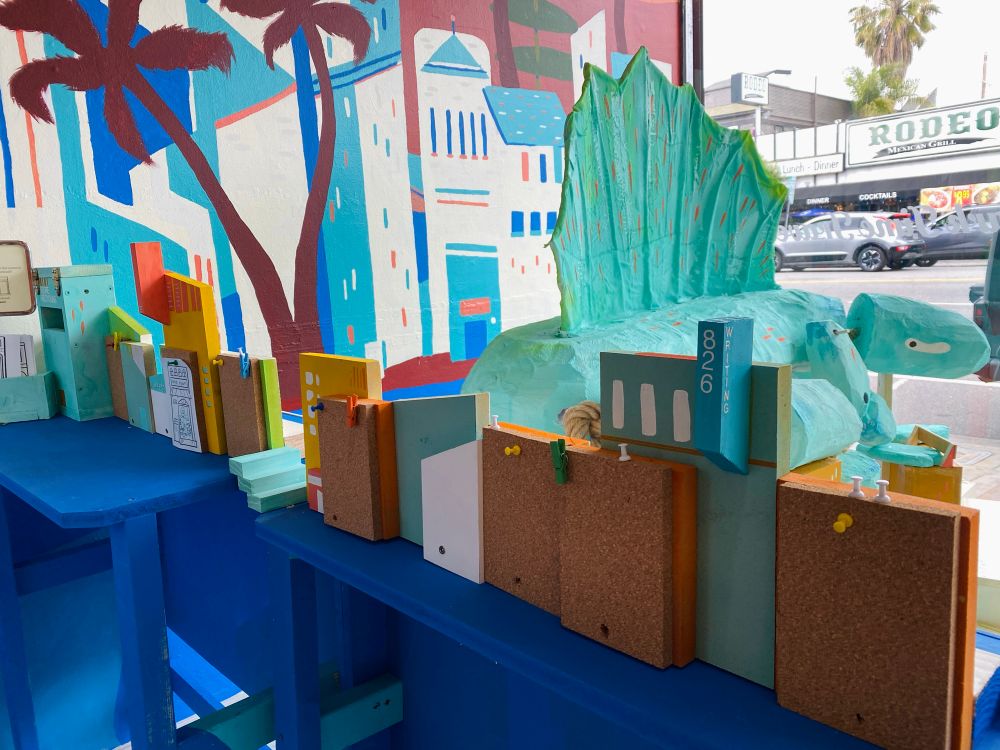 a storefront display of a prehistoric lizard resting on a bunch of blocky buildings. a small row of wooden buildings with corkboards are in the foreground.