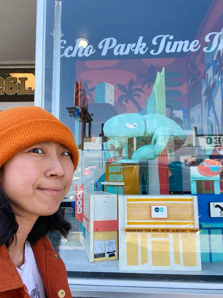 a person in an orange hat smiling in front of a store window which has a giant sculpture of a prehistoric lizard in it.