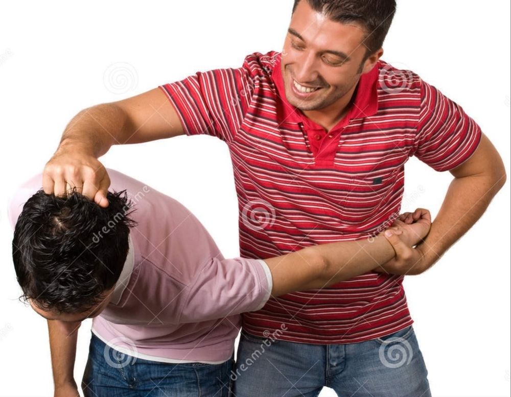 A brunet man in a striped red shirt and jeans holds another brunet man by his short hair, grinning as he forces him to bend at the waist while pulling the man's left arm behind him in a self-defense move.