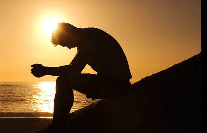 A man lit in silhouette looks down at the sunset, evincing a profound sense of grief