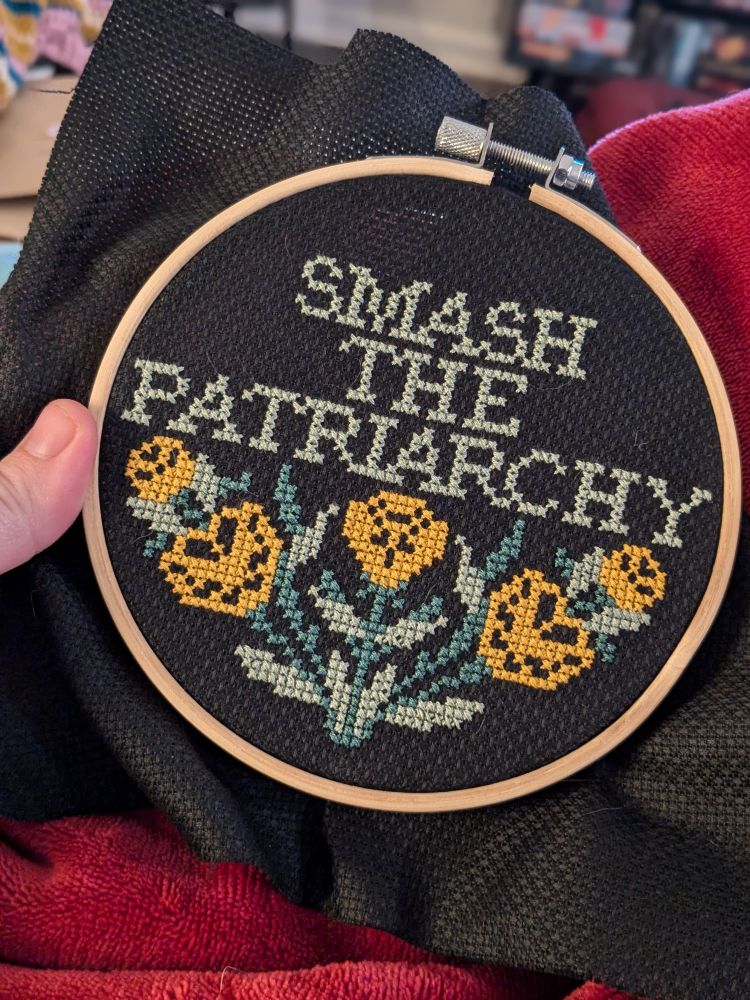 A photo of a cross stitch pattern in an embroidery hoop on black Aida fabric that says "smash the patriarchy" - 5 yellow flowers with green leaves and stems branch out beneath the text.