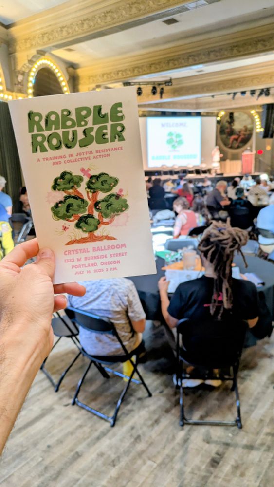 A photo of an event brochure being held up in front of a large room with many people seated around tables. 