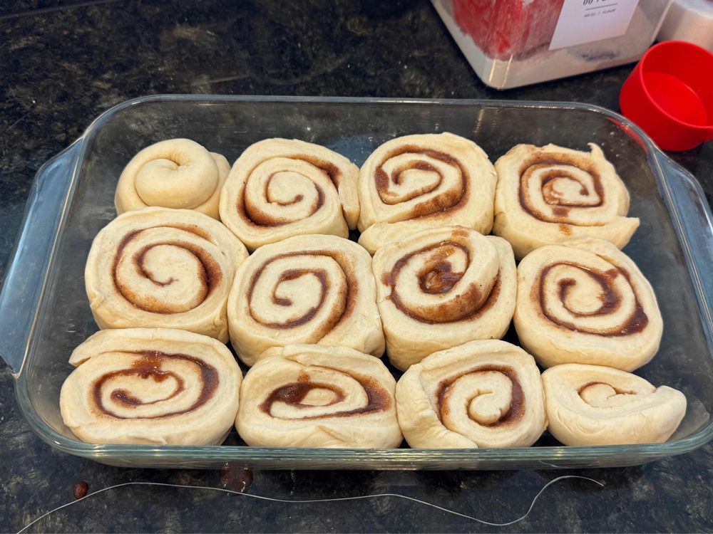 A glass dish containing a 3x4 collection of unrisen, unbaked pumpkin spice cinnamon buns, on a dark countertop with a bag of flour in the top-right.