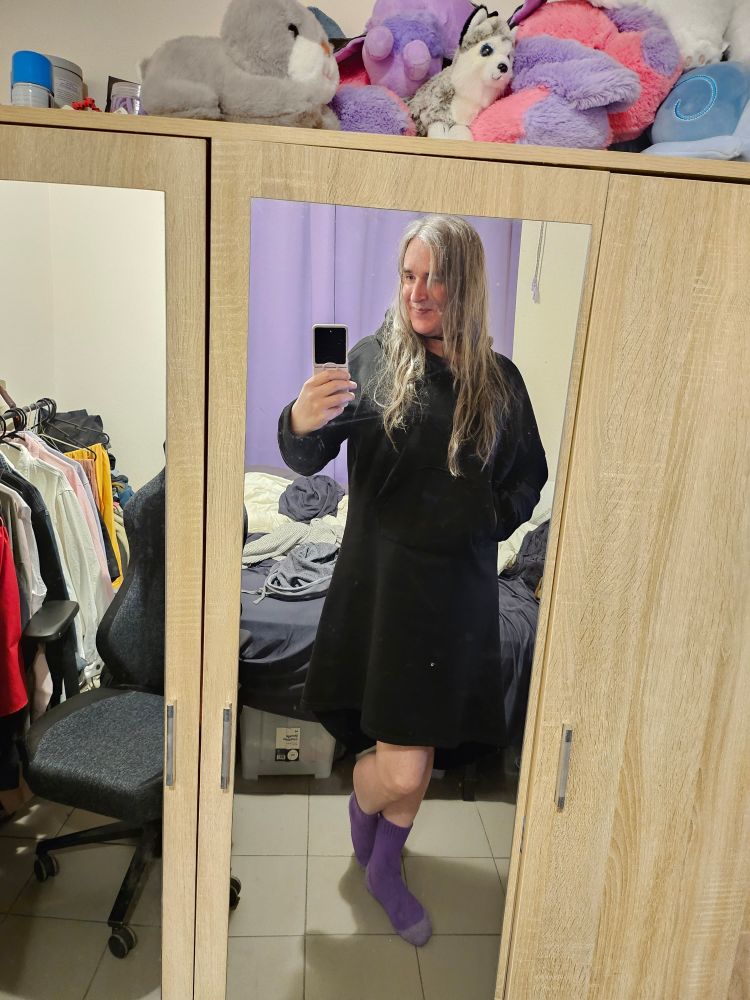 A girl in a hoodie dress and purple socks, standing in front of a mirrored wardrobe covered in plushies. Part of a chair and clothes rack in the other mirror