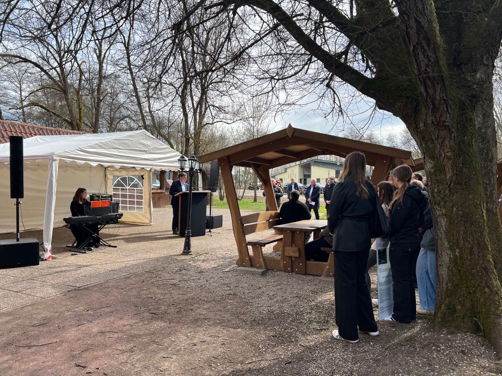 Andreas Froese hält die Festrede, auf der rechten Seite ein großer Baum, unter dem eine Gruppe von Schüler:innen steht, auf der linken Seite eine Musikerin am E-Piano