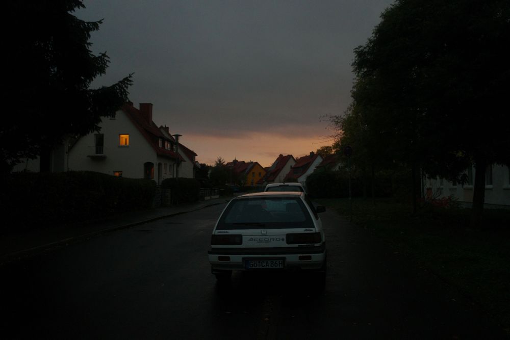 Photo of a cloudy sunset in a residential street. Cars and houses are visible.