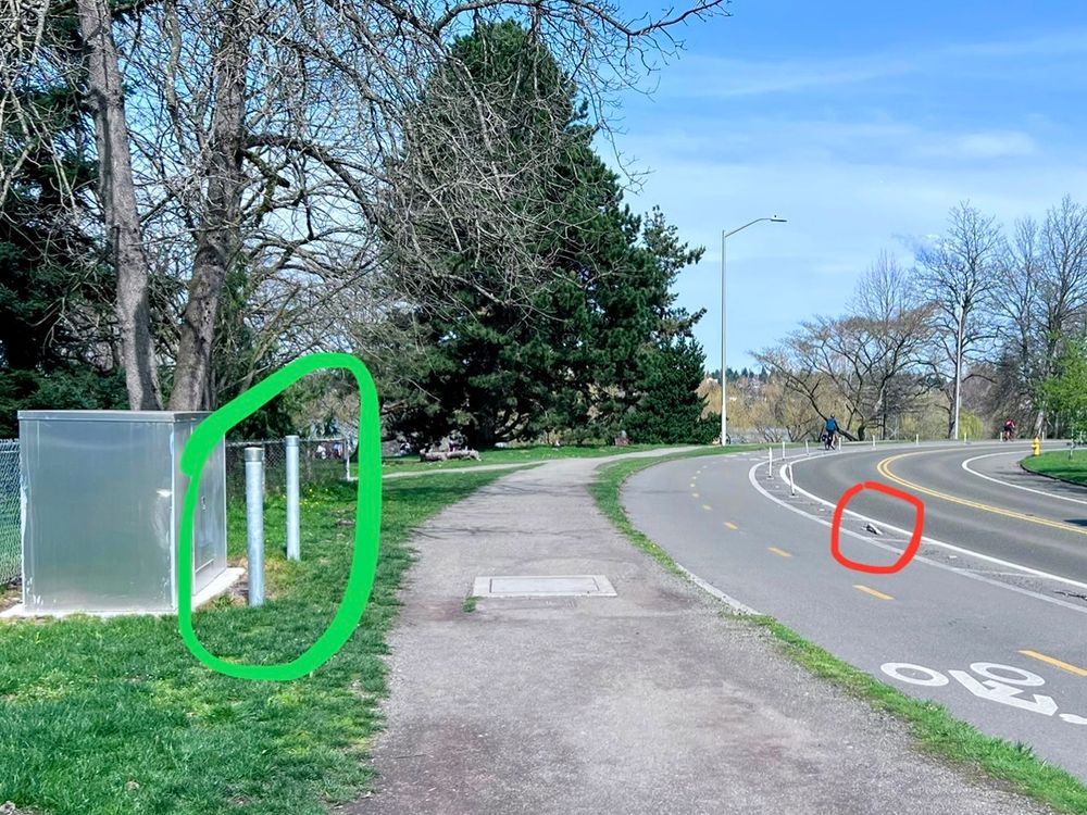 Bike lane around Green Lake protected by plastic flex posts (one is broken), while on the sidewalk a utility box is protected with metal bollard. That utility box was installed after the protected bike lane was installed.