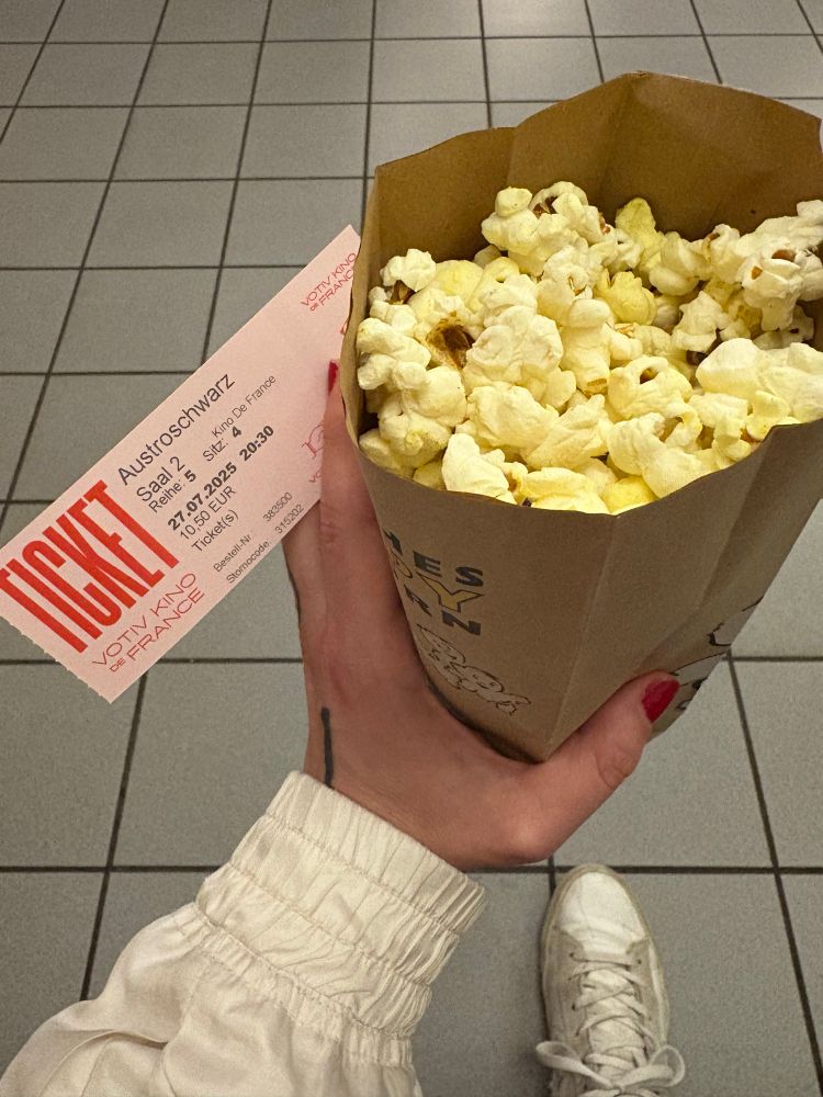 Man sieht aus der Perspektive der Person, wie sie ein Sackerl Popcorn in der Hand hält - und eine 
Kinokarte für den Film „Austroschwarz“ von Mwita Mataro