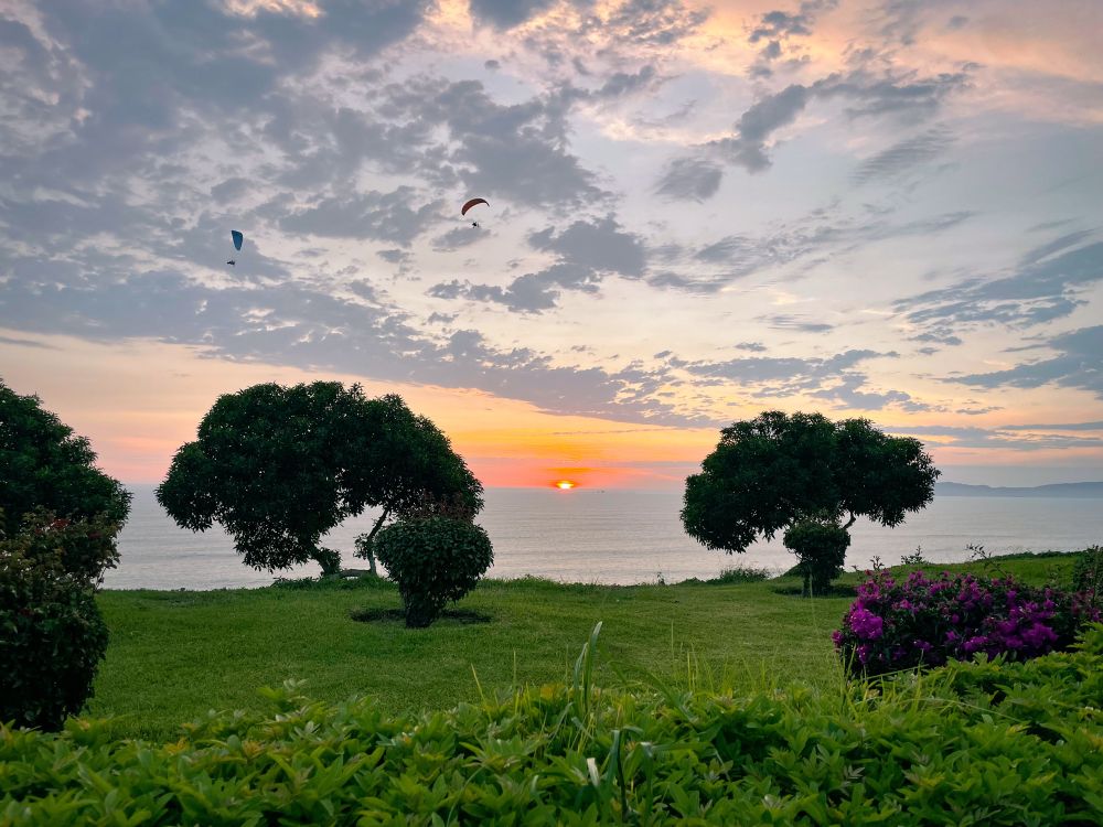 The sun sets over the Pacific Ocean in Lima, Peru. In the foreground are small trees and flower bushes, and in the background can be seen rocky islands. Two powered paragliders fly along the coast.