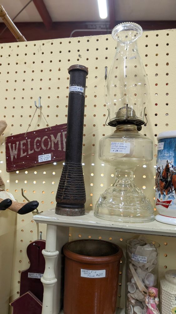 A wood rod labeled spool. The base is wider and ridged. There is also a welcome sign hanging to the left and a clear glass oil lamp to the right. 