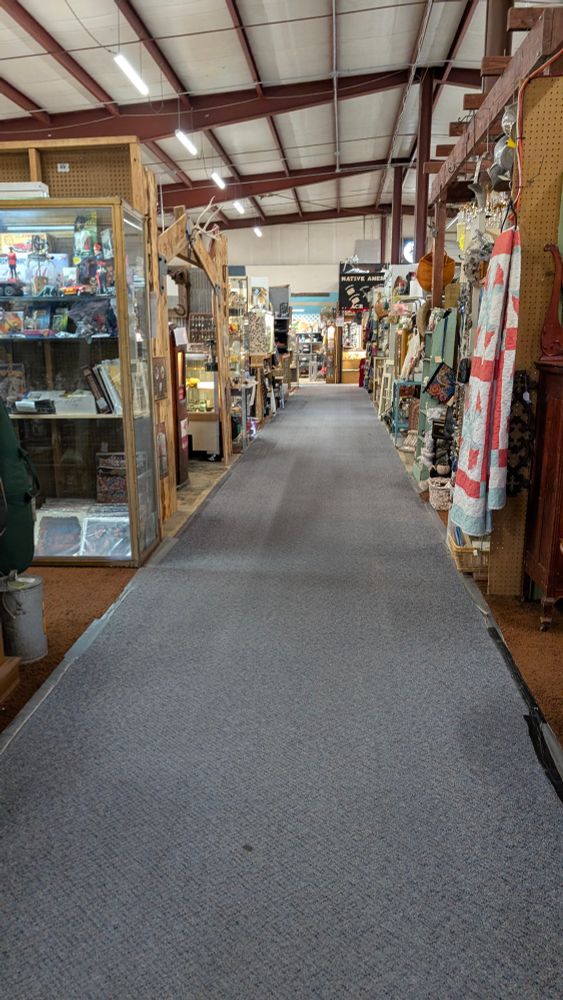 A gray carpet isle stretching before you set between shelves on either side, filled with miscellaneous items. The exposed rafters show that the building is a tall metal warehouse. 