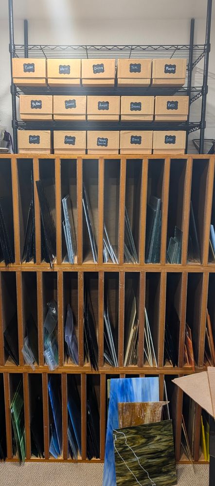 The bottom of the photo shows wood cabinets with tall but thin openings filled with sheets of glass standing upright. Above the wood cabinets is a black metal shelf with 3 rows of brown boxes. The boxes are 4x5x16 inches deep and have a small black label on the front indicating the color of scrap glass inside. 