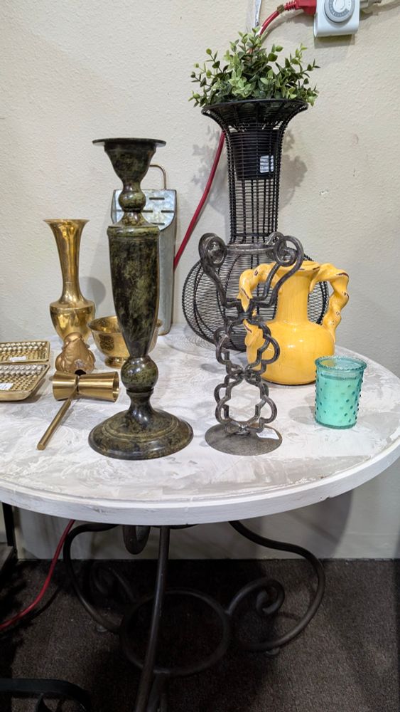 On the front of the white table is a green/black metal candle holder and to the right of that a gray metal candle holder that is bars bent into ornate shapes. Behind them is a collection of brass, black wire, yellow, and teal containers. 