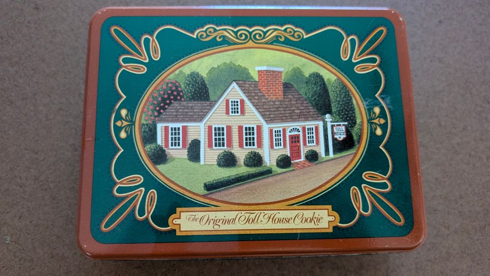 A green and brown tin with a framed image of a house on the lid. The text under the image says "The Original Toll House Cookie". 