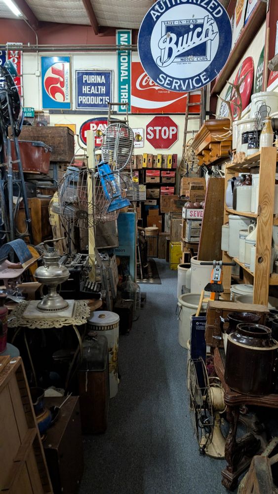 A tight path between cluttered shelves. The back wall has old wooden boxes and signs. The surrounding shelves are filled with ceramic jugs, old metal lamps, antlers, leather suitcases, and many other items. 