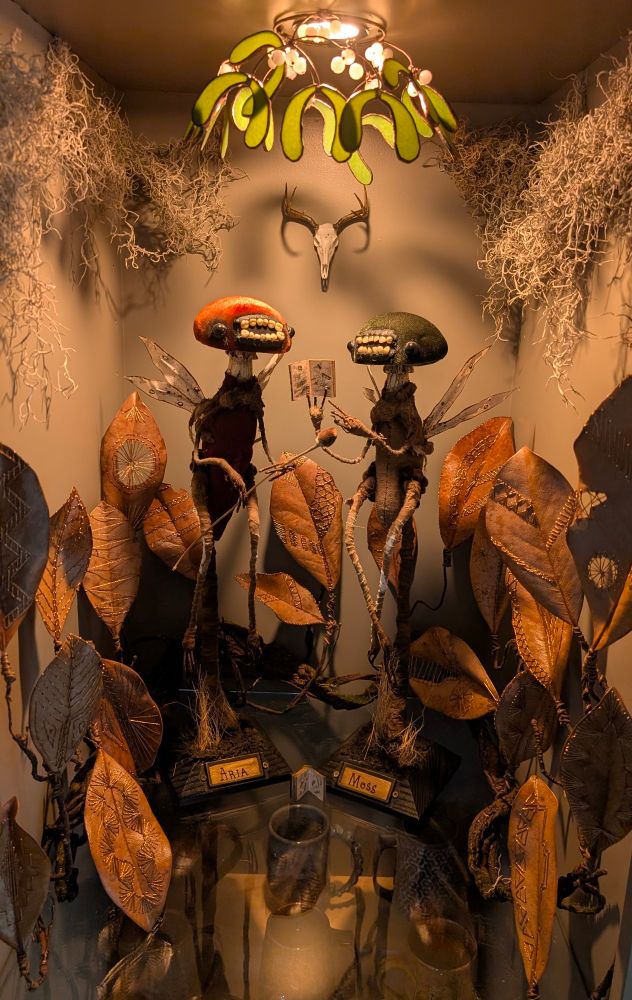 Two mushroom-headed tooth fairies (with very toothy-grins). The green one on the right is holding up a tiny book with insect illustrations. The orange one to the left seems very excited! There's a tiny deer skull hanging on the wall behind them with moss and mistletoe hanging from the ceiling. Copper-embroidered dead leaves surround them. A cozy environment for their studies...