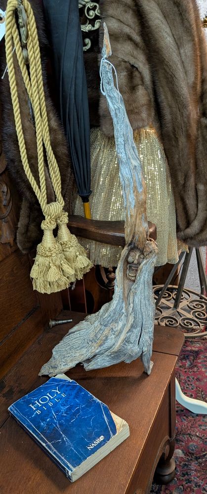 A piece of driftwood leaning against a fur coat. The piece of driftwood has an unhappy face carved into it. There is a bible placed near the base of the driftwood. 
