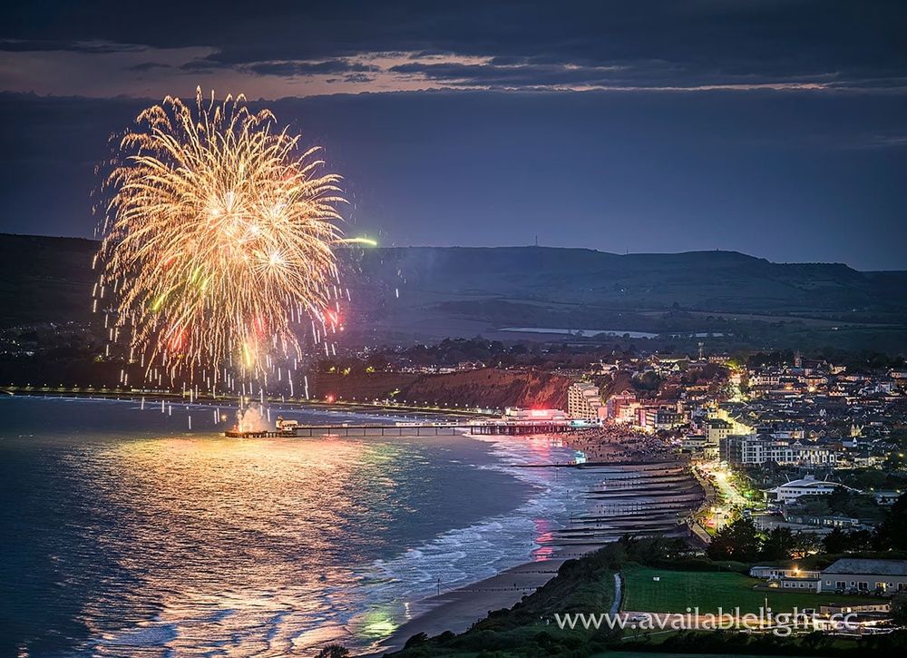 A huge gold firework above a pier. A coastal town is lit at nightfall. Reflections are on the wet sand and the sea.