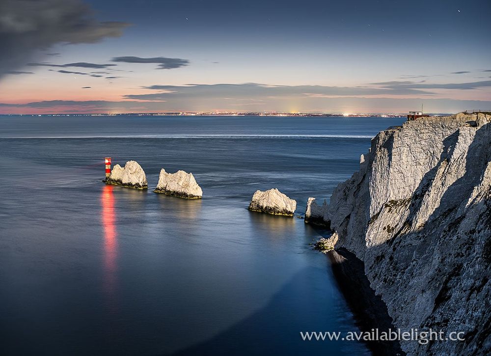 Night falls over a chalk cliff and rocks. A red and white lighthouse guards the rocks. Moonlight illuminates the scene.