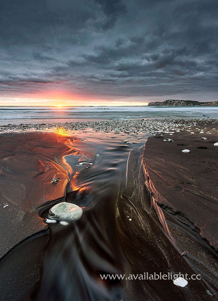 The sun about to dip below the horizon over the sea. The camera is looking down a water inlet in the sand, white pebbles scattered around. A chalk headland is to the left of the image. The golden light of the sunset is reflecting on the wet sand.