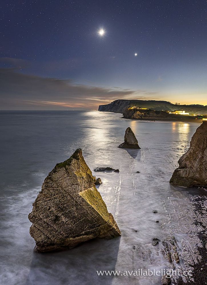 It's almost dark, the Moon and Venus prominent in the sky. A calm sea surrounding 2 large rock stacks. A cliff and coastline complete the scene.