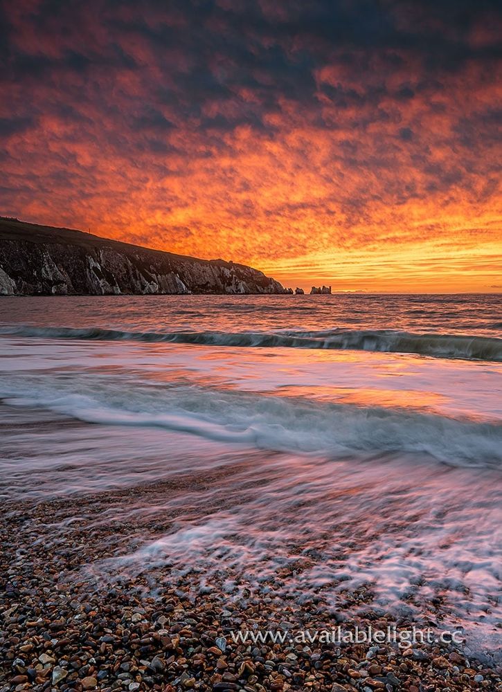 A fiery sunset over a beach. Vivid orange and yellow fills the sky. A chalk headland in the distance with jagged stacks and a lighthouse at the end. The tide rolls into the shore, and recedes over the pebbles. 