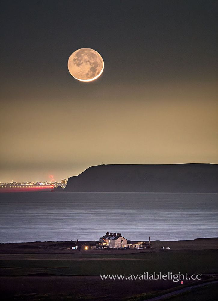 A large moon, in an almost dark sky with the illumination of a distant city. Several coastguard cottages are closer in the view, a headland and a lighthouse in the middle space.