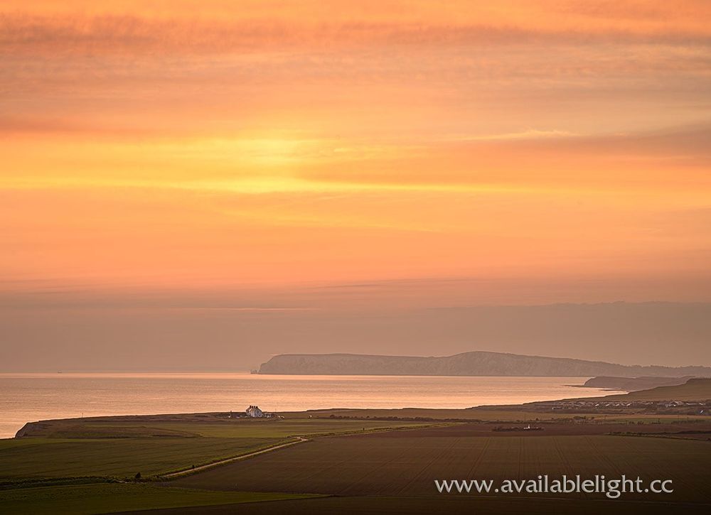 An orange and yellow sunset dominating a wide view of a coastal road and headland.