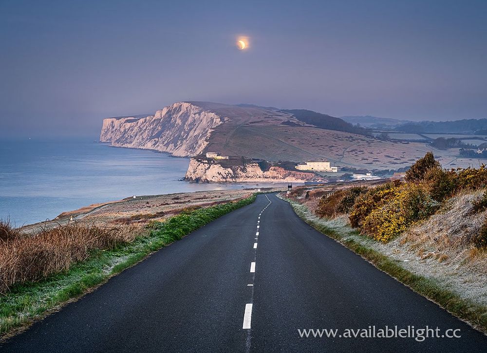 A coastal road, the moon hazy in the sky as dawn breaks. A chalk headland lies beyond the scenic road.