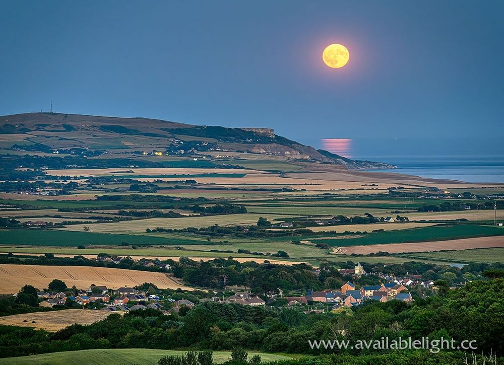 The moon rising above coastal farmland, a village in the foreground. The sea is reflecting the moon.