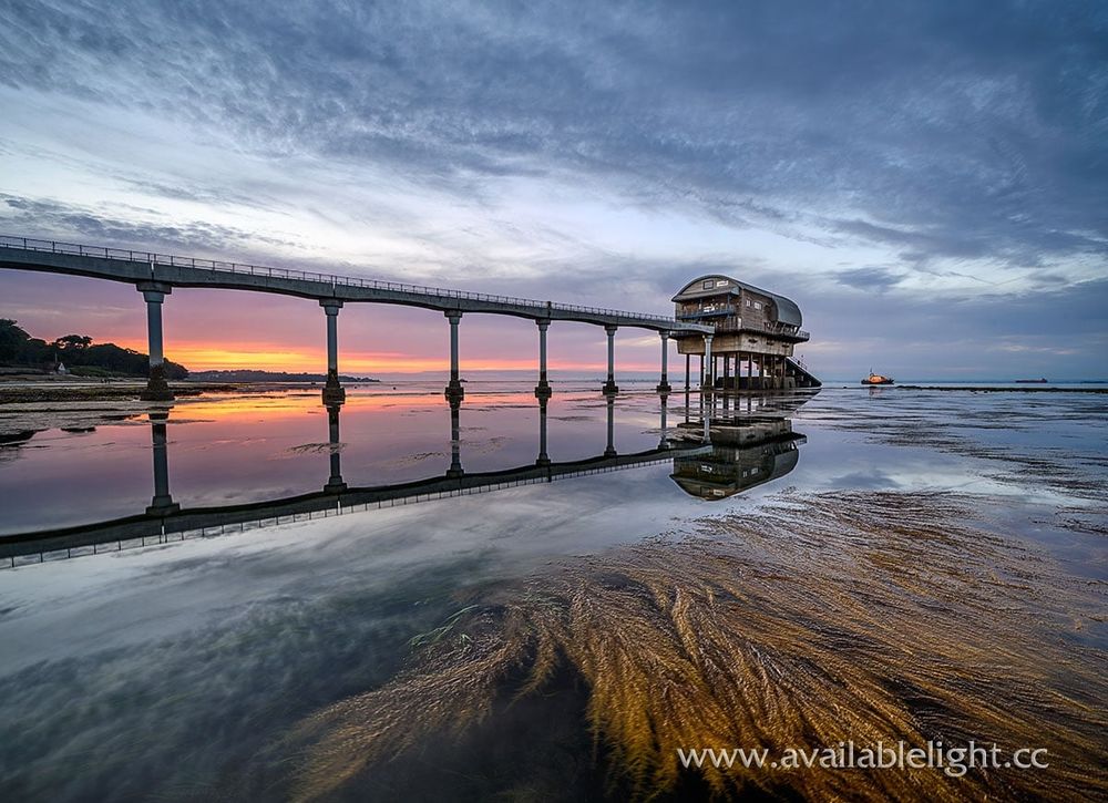 Gentle pinks, yellows and orange in a grey blue sky over a lifeboat station at low tide. Seaweed is visible in the rock pools and the lifeboat is moored in the distance.