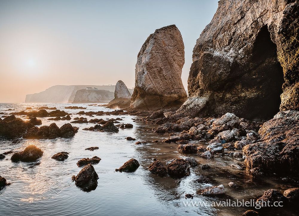 A rocky beach, caves, large rock stacks and a distant headland, the sun is low in the sky.