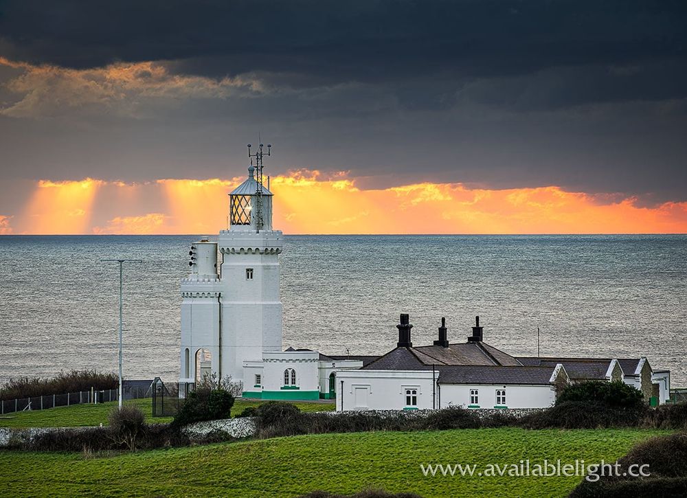 A white lighthouse and cottages sit in a field next to a steely calm sea. The sun is about to emerge from a low cloudbank just before it sets. The sky is overcast with cloud but an orange and yellow glow fills the horizon.