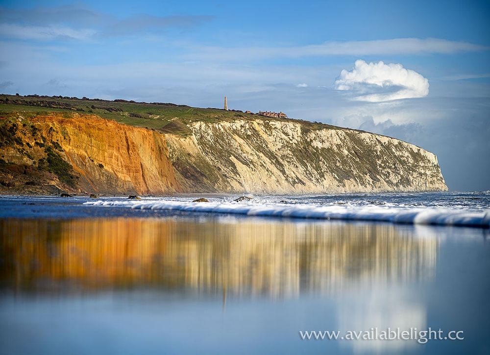 A chalk and clay cliff is reflected in wet sand as the sea rolls in. The tide is rough on a stormy day, but the sky is blue.