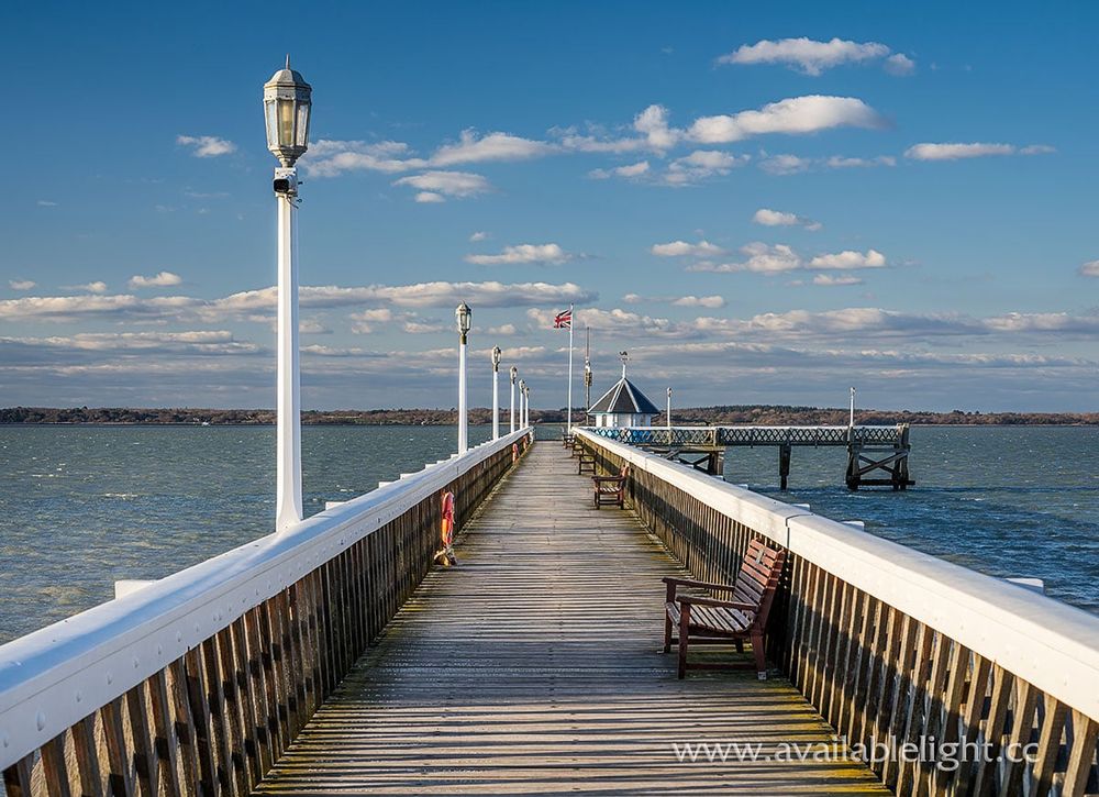 A white railed pier, the view leading down to the end with a hut, flag , several benches and ornamental lamposts. The sky is blue with a few white fluffy clouds. The sea is calm but with a few waves rippling alongside the pier.