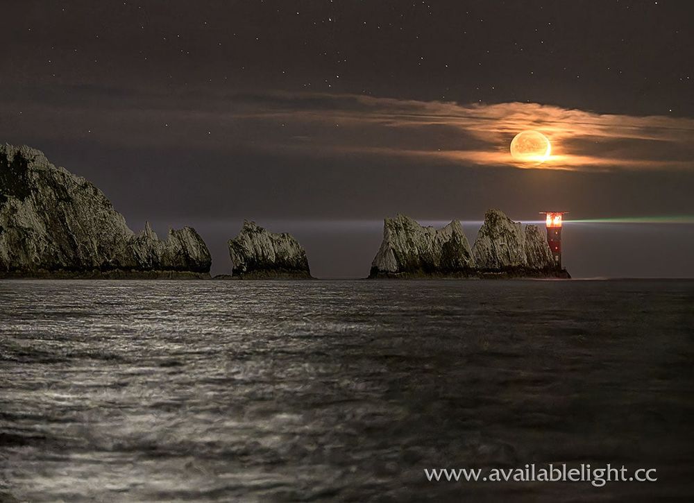 A very dark sky and inky sea. The moon is low over a lighthouse which is casting it's beam across the centre of the photo. The red and white lighthouse stands at the end of jagged chalk stacks.