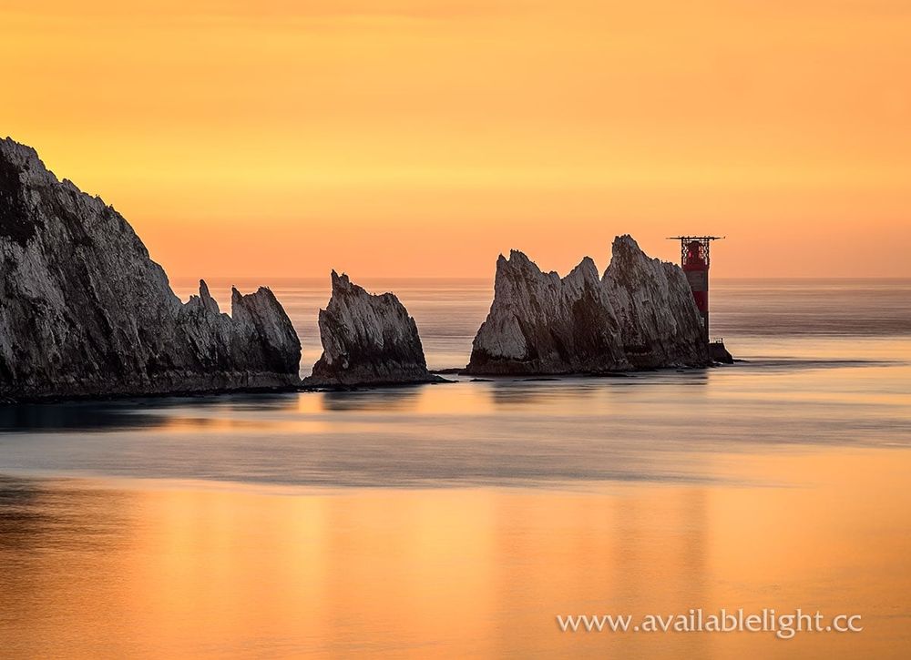 A fiery orange and yellow sky, reflecting in a calm sea which is surrounding chalk stacks and a red and white lighthouse.