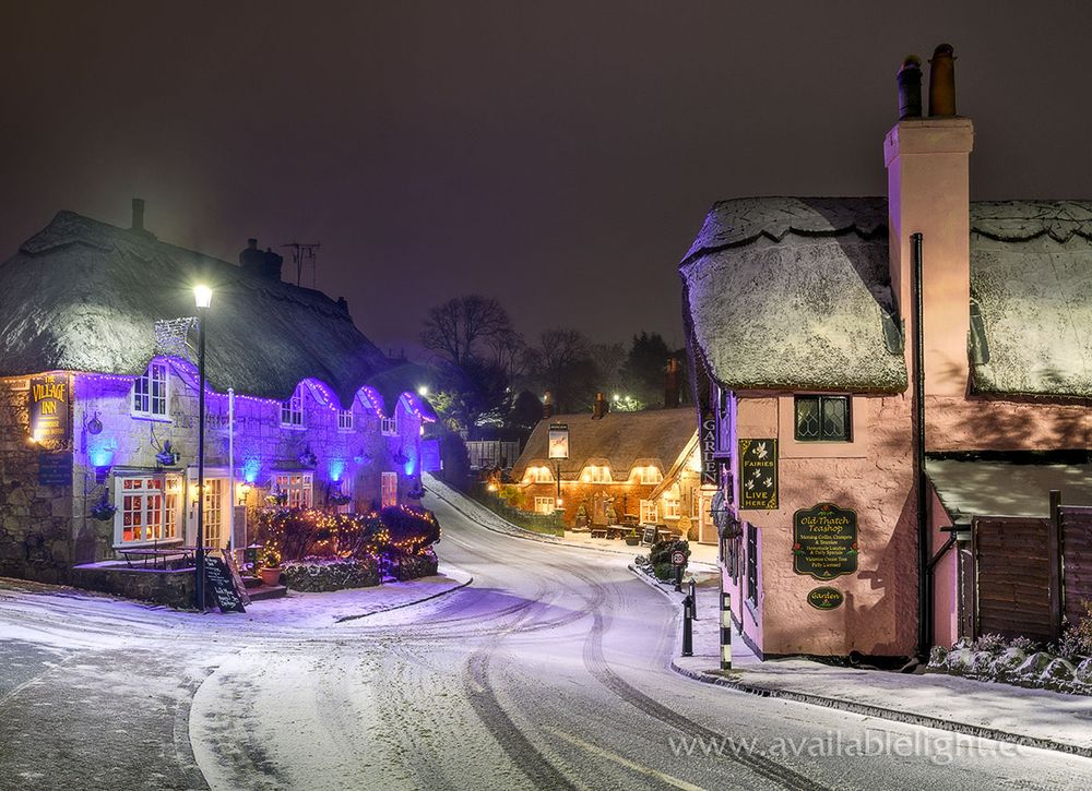 A light dusting of snow covers several thatched buildings. A small road winds between them, artificial lights illuminate this night time quaint winter scene.